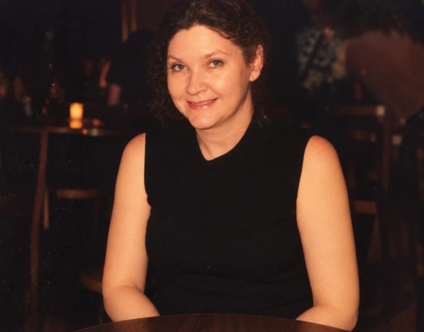 Francis Ann Dillard Hanna in a black sleeveless top at a restaurant table, smiling, candlelit tables behind her